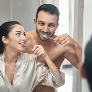 Attractive couple brushing teeth in the mirror, symbolizing the healthy lifestyles supported by Derby Dental Care.